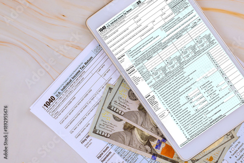 Digital tablet displays tax 1040 forms while US cash banknote is scattered on table with other tax documents.