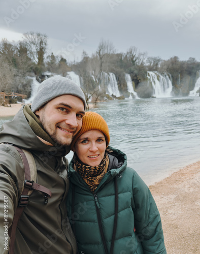 Couple posing for a selfie near Kravica Waterfall in Bosnia and Herzegovina, with wide cascades, river water, misty air, and natural landscape creating a calm travel atmosphere.