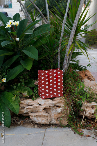 Red paper bag with hearts  on a stone ledge surrounded by lush tropical plants and flowers.
