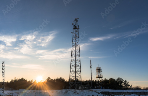 Tall telecommunication tower with radar and antennas against a blue sky at sunset. Winter landscape with snow, forest silhouette, and bright sunburst flare through the trees.