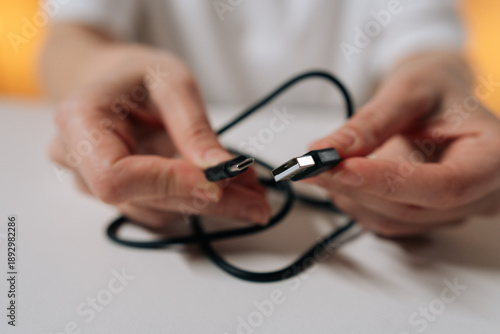 Photos Close-up hands of unrecognizable person untangling knotted black power cord sitting at white table, preparing charging cable for use and restoring order to tangled wire