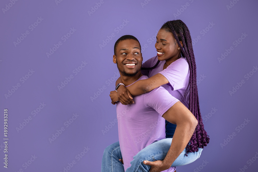 Obraz premium Piggyback Ride. Smiling black man carrying and riding his woman with long afro braids on back, enjoying time together, standing isolated on violet studio background, having fun, free copy space