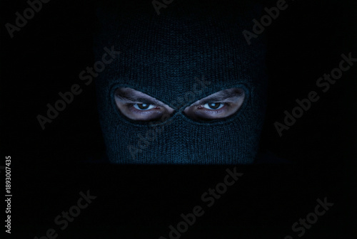 A close-up shot of a person in a black ski mask with intense eyes peering out from the darkness. The person is shrouded in shadow, with only their eyes and the mask visible.