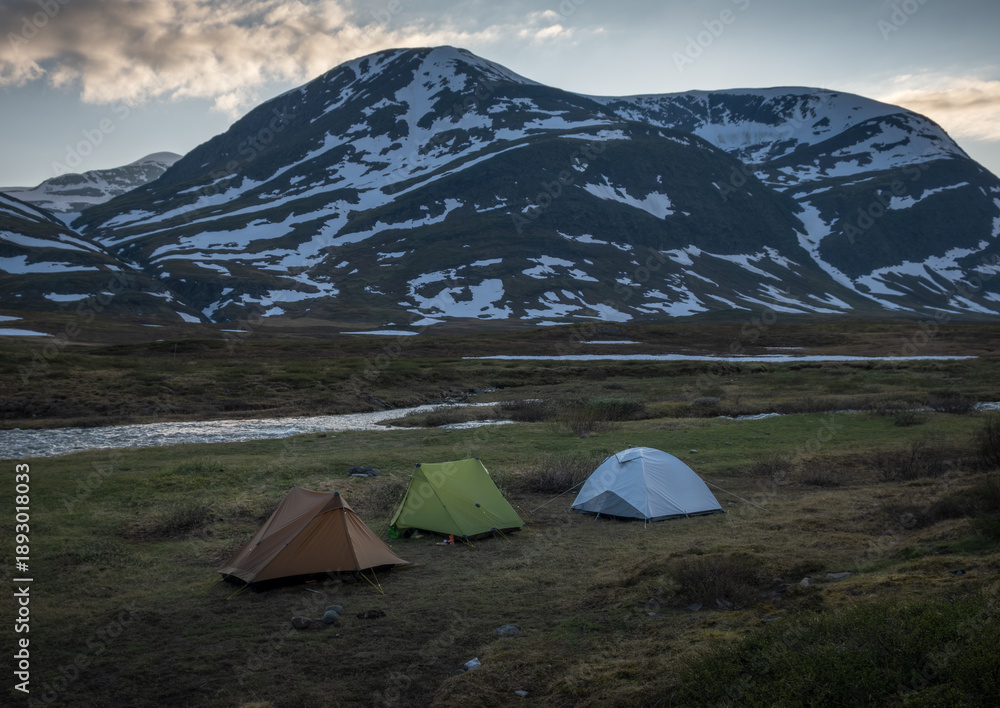 Obraz premium Three colorful tents stand on a grassy riverbank in Swedish Lapland, with snow patched mountains rising in the background along the Kungsleden trail