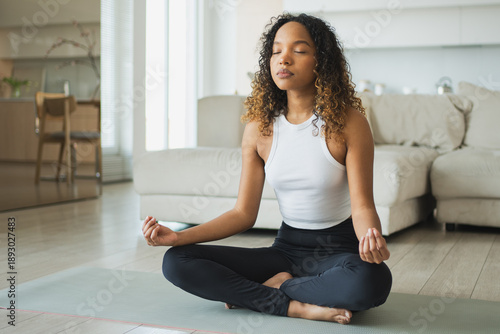 Yoga mindfulness meditation. Young healthy African girl practicing yoga at home. Woman sitting in lotus pose on yoga mat meditating relaxing indoor. Girl doing breathing practice. Yoga at home