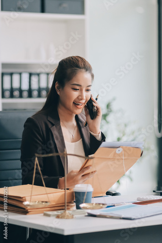 Confident Lawyer Working at Desk: A young Asian female lawyer confidently works on her laptop, surrounded by legal symbols. She exudes professionalism and competence in her modern office setting.