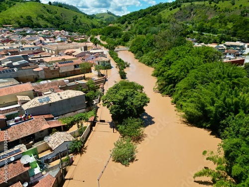 Vista aérea por drone de uma enchente urbana, mostrando ruas alagadas, casas parcialmente submersas e o impacto das chuvas intensas. Conceito de desastre natural, mudanças climáticas, crise ambiental.