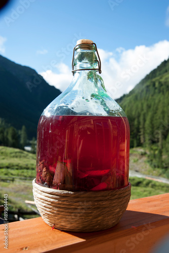 Tyrolean Swiss stone pine liqueur (Zirbala) infusing in glass demijohn bottle on sunny alpine balcony, mountain valley background