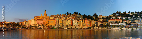 Panoramic sunrise view of Menton, France, with colorful old town buildings glowing in warm light, set against the Mediterranean Sea and blue sky on the French Riviera