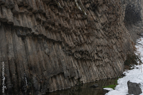 Vertical basalt columns rise above narrow river at Symphony of Stones, dark volcanic rock textures contrast with snow, flowing water in winter light
