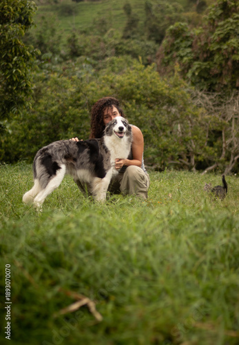 Happy woman crouching and hugging her Australian Shepherd or Border Collie dog in a rural meadow. A small tabby cat walks in the grassy background. Pet owner lifestyle concept with multiple animals.