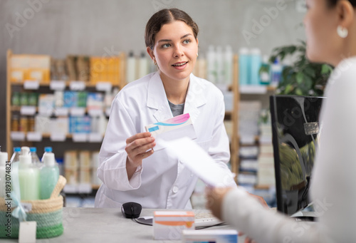 Professional young pharmacist using computer to check medication stock, politely explaining treatment options, and processing order for female customer