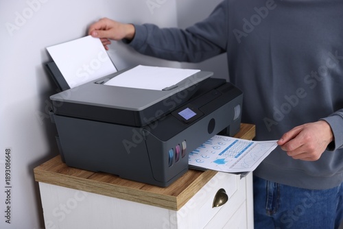 Obraz na plátne Man taking printed sheet of paper from printer at chest of drawers near grey wal