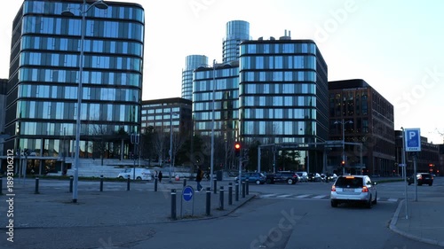 A view of a modern building in the capital of Denmark. Lots of traffic and people walking around the city. Large windows on the buildings