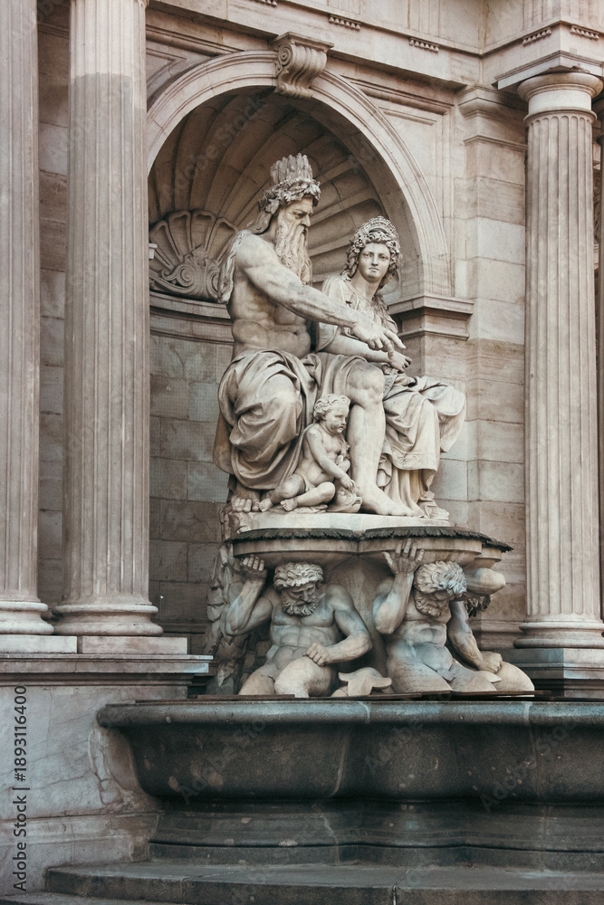 Obraz premium Detailed view of an ornate Baroque fountain sculpture in Vienna, Austria, featuring classical mythological figures carved in stone against a grand architectural facade with columns and arches. 