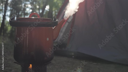 The fire from the stove in the camping site with the coffee pot on the top and the tent on the back. A holiday theme in nature. Defocused close-up shot.