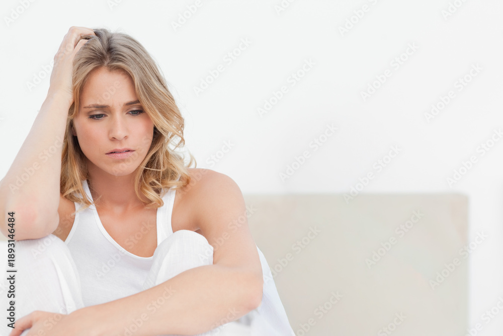 Obraz premium Woman wearing white tank and light pants, sitting on beige-backrest sofa, looking down, copy space