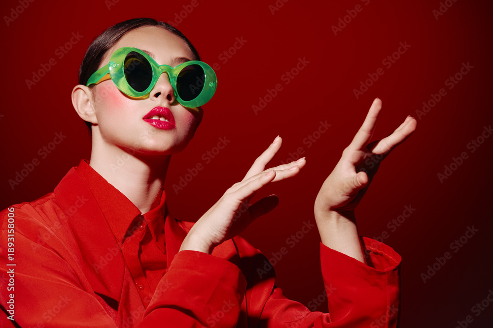 Fototapeta premium Fashion model with bold green eyewear and bright red lipstick poses with expressive hands wearing a matching red outfit against a red background. Creative beauty concept.