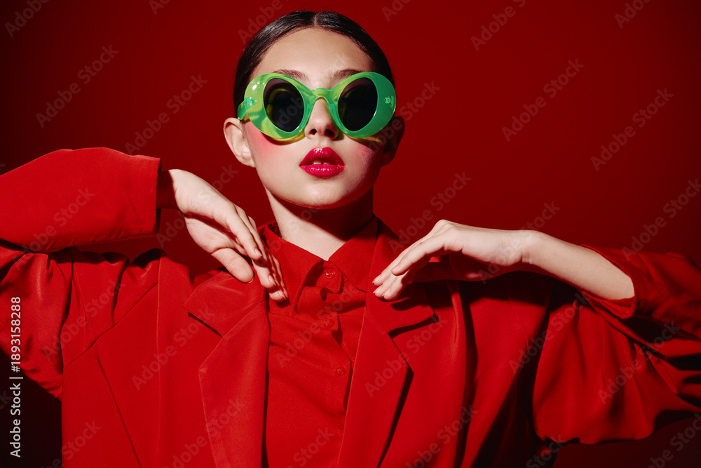 Fototapeta premium Young woman in oversized green sunglasses wears vivid red coat and lipstick posing stylishly against a red background fashion fashion beauty creative portrait studio modern.