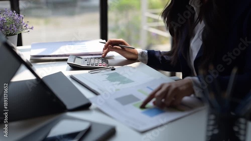 A woman is sitting at a desk with a calculator and a piece of paper with graphs on it. She is pointing at the paper and she is focused on the numbers