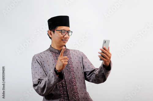 An Asian Muslim man smiles happily while sending a message using his mobile phone, isolated on white background