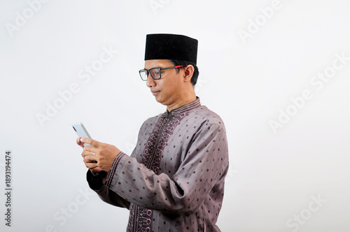 An Asian Muslim man smiles happily while sending a message using his mobile phone, isolated on white background