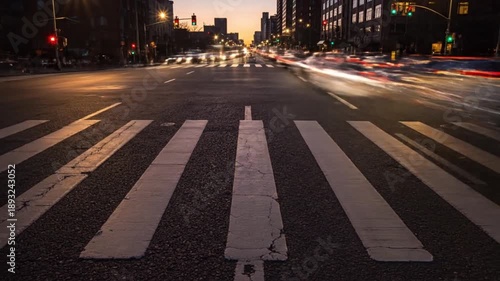 A city street at dusk with a crosswalk in the foreground and blurred traffic moving through the background.