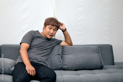 Wallpaper Mural Asian man sitting casually on a modern grey sofa in minimal indoor space. His relaxed posture represents comfort, home lifestyle, and a calm, stress-free environment Torontodigital.ca