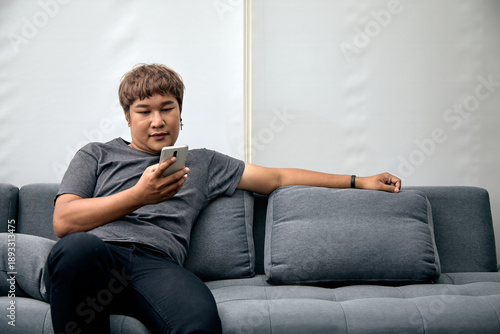 Wallpaper Mural Asian man relaxing on sofa while using smartphone in modern indoor setting. Casual lifestyle moment reflecting comfort, technology use, and digital connectivity at home Torontodigital.ca