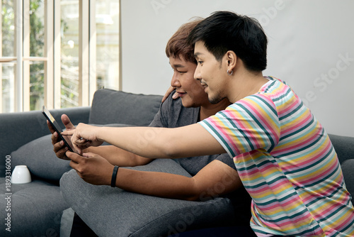 Wallpaper Mural Two Asian men sitting close together on sofa using digital tablet. Their shared focus and joyful expressions reflect friendship, digital bonding, and modern lifestyle at home Torontodigital.ca