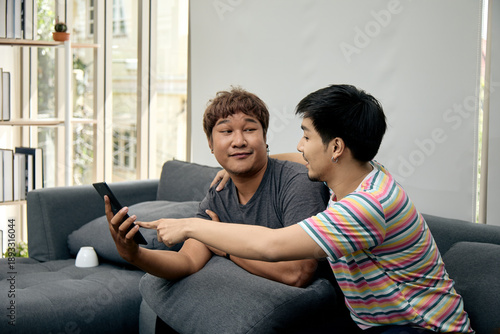 Wallpaper Mural Two Asian LGBTQ men sitting close on sofa, smiling and pointing at a tablet while making eye contact. The moment reflects fun interaction, friendship, and digital lifestyle at home Torontodigital.ca