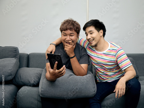 Wallpaper Mural Two LGBTQ Asian men sitting on sofa, smiling and enjoying watching content on tablet together. This moment reflects digital bonding, friendship, and relaxed lifestyle at home Torontodigital.ca