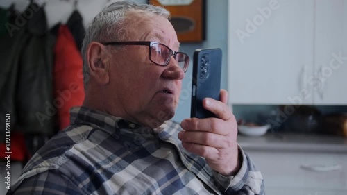 Close-up of elderly man in glasses using his smartphone to record voice message while sitting in cozy home kitchen. Modern technology, communication, and elderly people adapting to digital life.