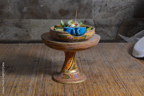 An ornate Dulang pedestal holding a ceramic bowl with blue cups and ritual offerings sits on a woven bamboo surface, illuminated by soft artificial lighting in a Balinese home at night.