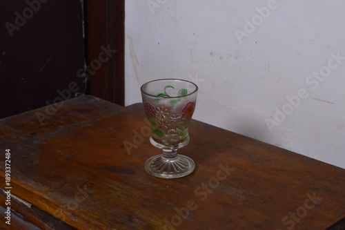 Perspective view of a single vintage clear glass goblet featuring colorful embossed flower designs, placed on a dark wooden table against a plain white wall in indoor night lighting.