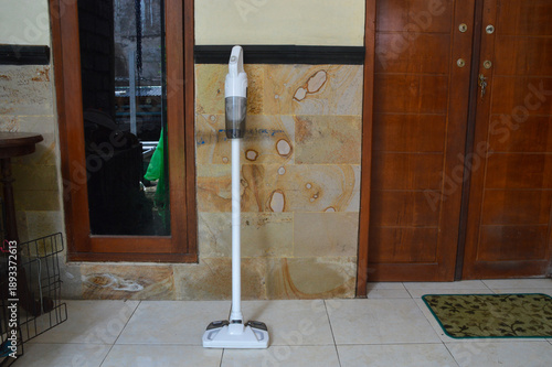 A sleek white battery-powered vacuum and mop appliance is placed upright against a textured sandstone wall on a tiled porch floor between a window and a wooden door.