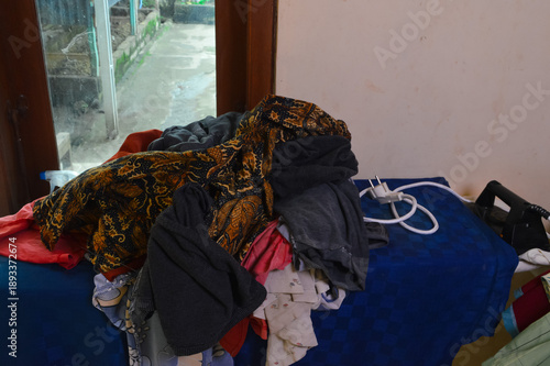 Various colorful clothes and a traditional batik fabric sit in a heap on a blue checkered ironing board next to an electric iron and a window during the day in a domestic setting.