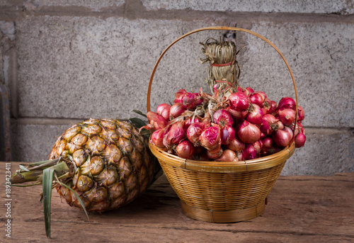 Tableau sur toile Red onions in wicker basket and fresh pineapple on wooden table with concrete wall background