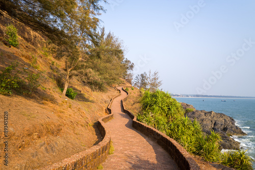 Curved stone walkway hugs the hillside above rocky cliffs and overlooks the Arabian Sea at Bekal Fort, India. Warm sunlight casts gentle shadows on the textured path and surrounding greenery.