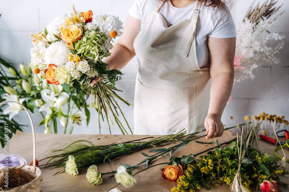 Naklejka premium Hands of florist hold large floral bouquet over workbench in studio. Sustainable wedding decor, micro-wedding aesthetics, floral symbolism.