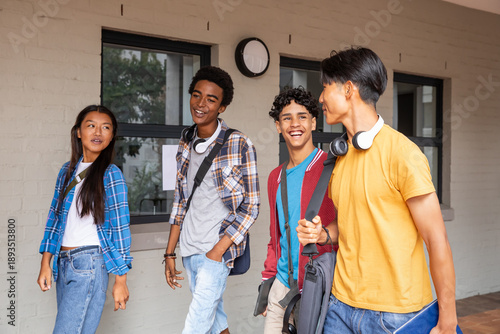 Diverse student friends walking and talking along campus corridor with backpacks and tablet
