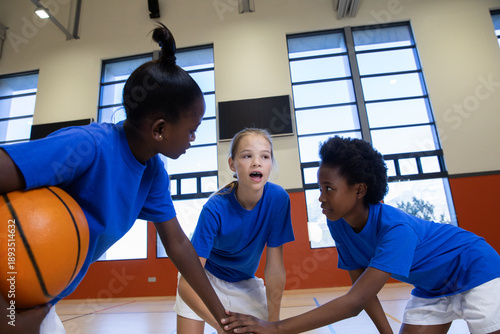 Diverse youth basketball teammates huddling with hands on basketball on gym court