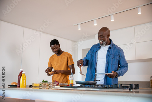 African american father and son cooking on kitchen island with spatula, stainless steel frying pan