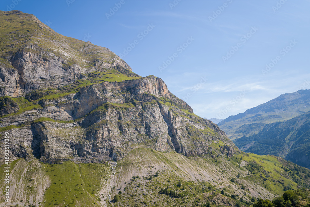 Obraz premium Jagged rock faces and grassy ledges rise sharply under a clear blue sky in the mountains near Gavarnie Gedre, French Pyrenees. The landscape features dramatic elevation, rugged textures, and summer