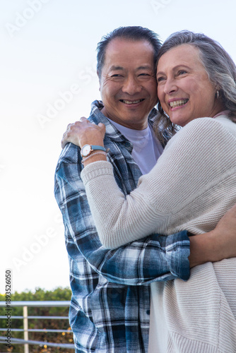 Diverse senior couple embracing on balcony with metal railing showing wristwatch, bracelet