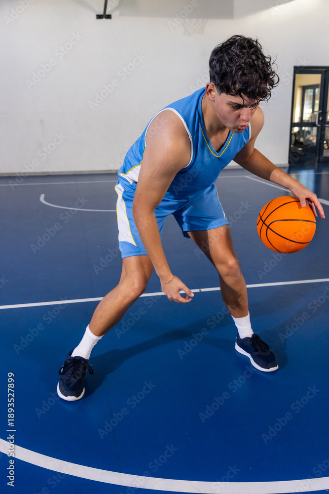 Fototapeta premium Teenage male athlete dribbling orange basketball on court in gym near hoop, glass-panel door