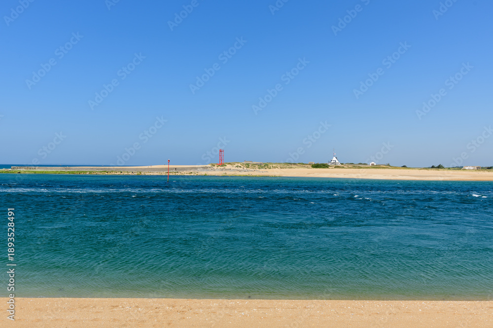 Fototapeta premium Vivid blue tidal water flows along a wide sandy shoreline under a clear sky at Barre d'Etel in Brittany. Distant navigation markers and a white signal tower add structure to the bright, tranquil