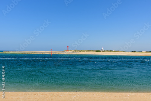Vivid blue tidal water flows along a wide sandy shoreline under a clear sky at Barre d'Etel in Brittany. Distant navigation markers and a white signal tower add structure to the bright, tranquil