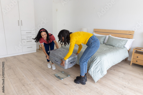 Diverse mom and teenage daughter lifting gray suitcase onto digital luggage scale in bedroom