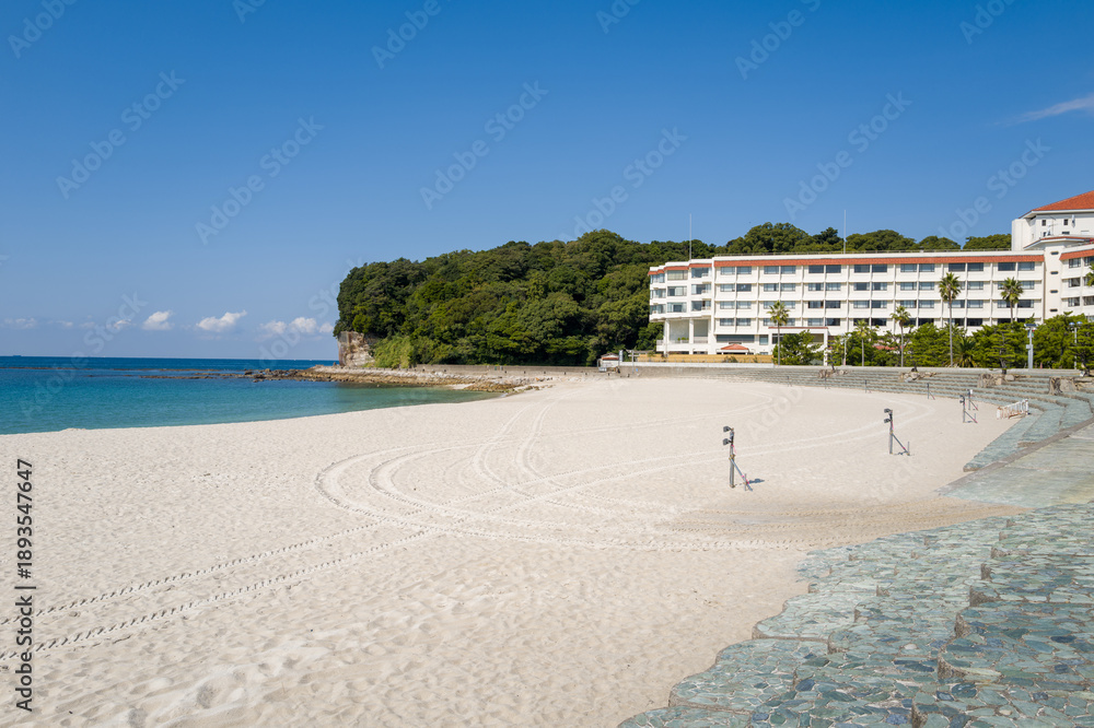 Obraz premium Wide expanse of pristine white sand borders turquoise water, with a modern beachfront hotel and lush green trees under a clear blue sky in Shirarahama, Japan.